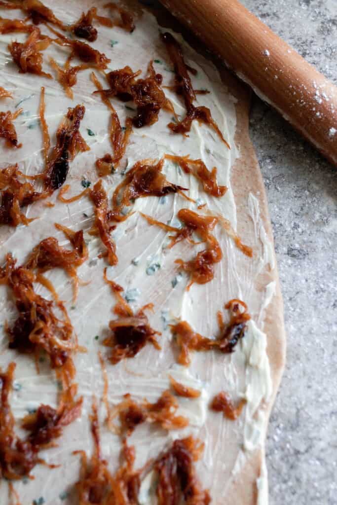 sourdough roll dough rolled out on a countertop with herb butter and caramelized onions spread out.