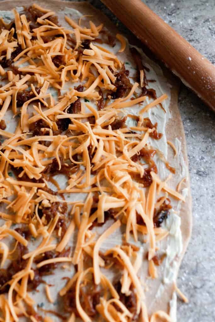 sourdough rolls dough rolled out with herbed butter, caramelized onions, and shredded cheese on the dough