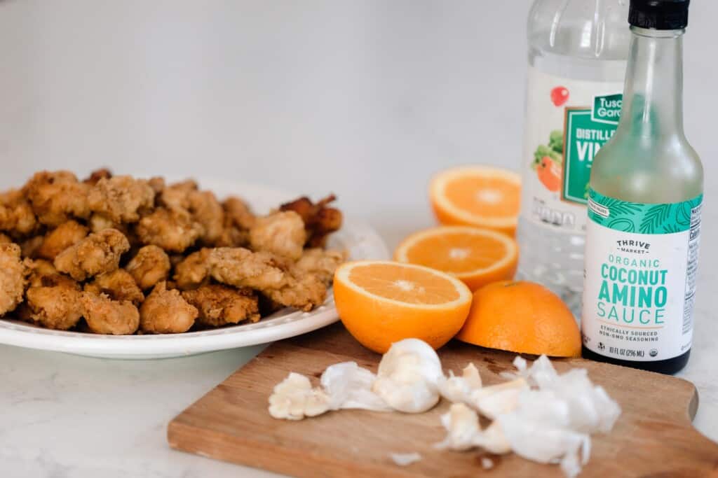 einkorn fried chicken on a white plate with oranges sliced in half, a cutting board with garlic on top. A bottle of coconut aminos and vinegar sit behind the cutting board.