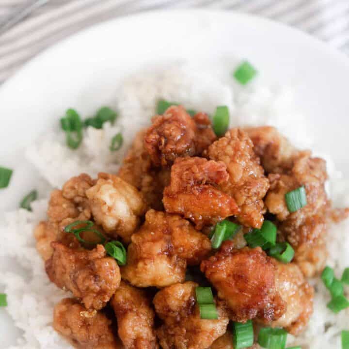 overhead photo of homemade orange chicken on top of white rice and topped with green onions on a white plate with a gray and white napkin in the background