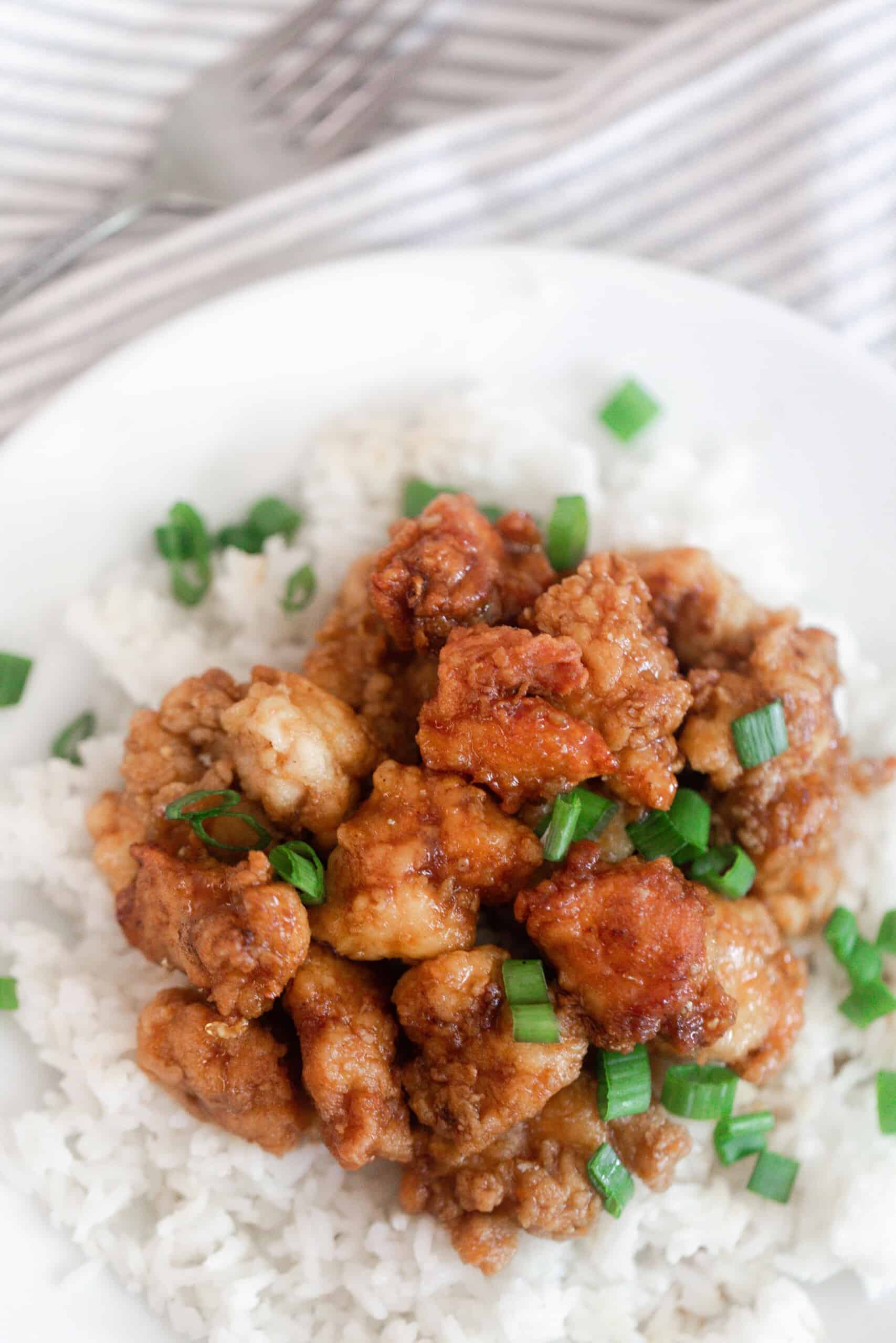 overhead photo of homemade orange chicken on top of white rice and topped with green onions on a white plate with a gray and white napkin in the background