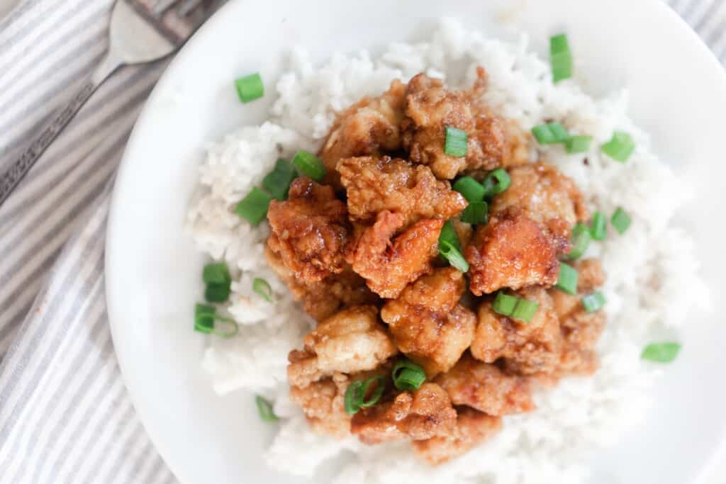 overhead photo of homemade crispy orange chicken made with einkorn flour on top of white rice with sliced green onions sprinkled on top. The plate sits on top of a gray and white stripped towel with a fork to the left