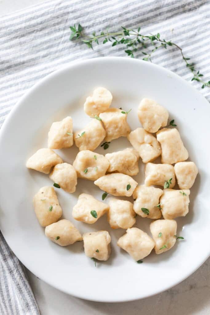 overhead photo of a white bowl full of homemade gnocchi with fresh herbs sprinkled on top. a white and blue stripped towel topped with fresh thyme sits behind the bowl.