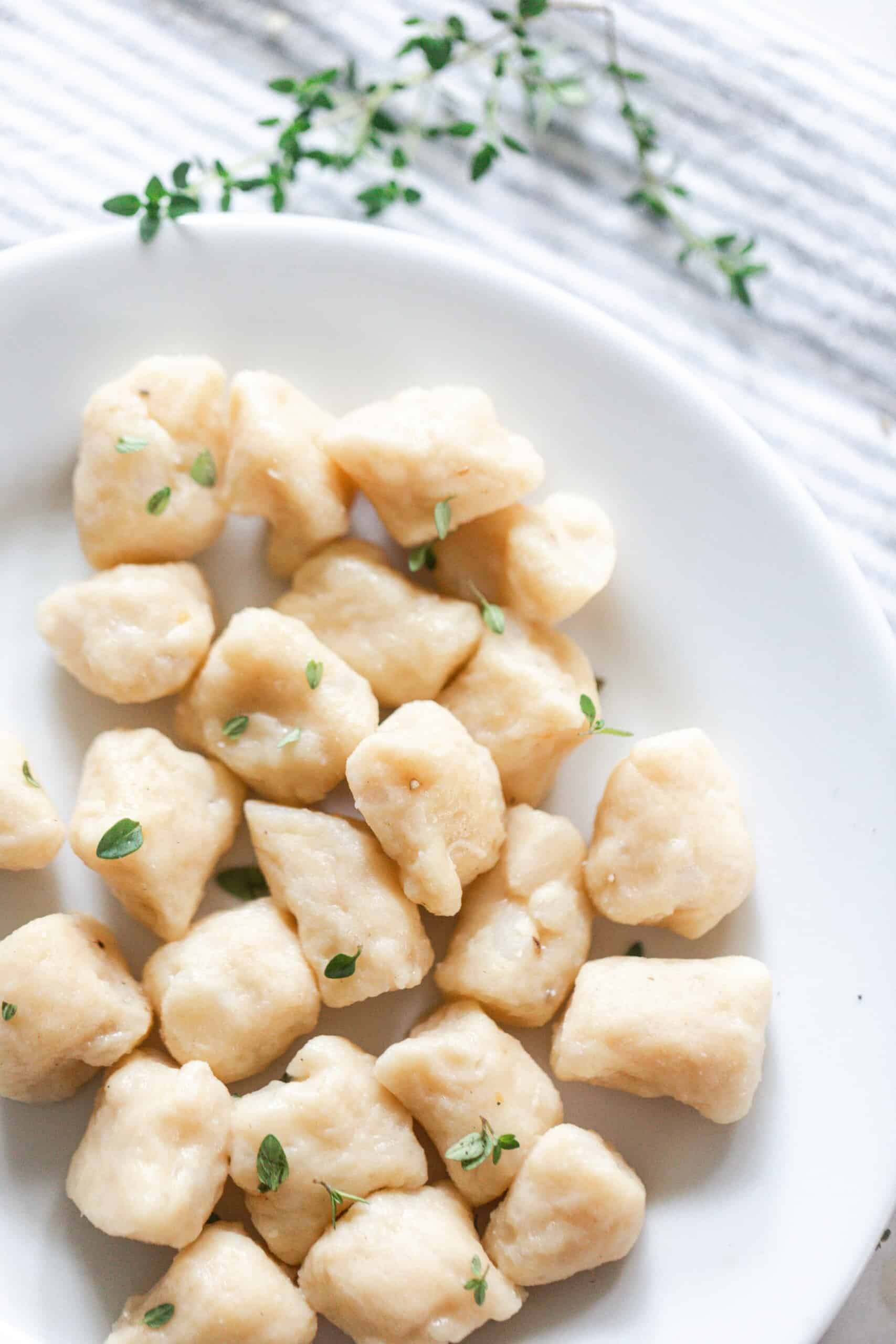 overhead photo of homemade gnocchi in a white bowl with thyme on top of a blue and white stripped napkin in the background