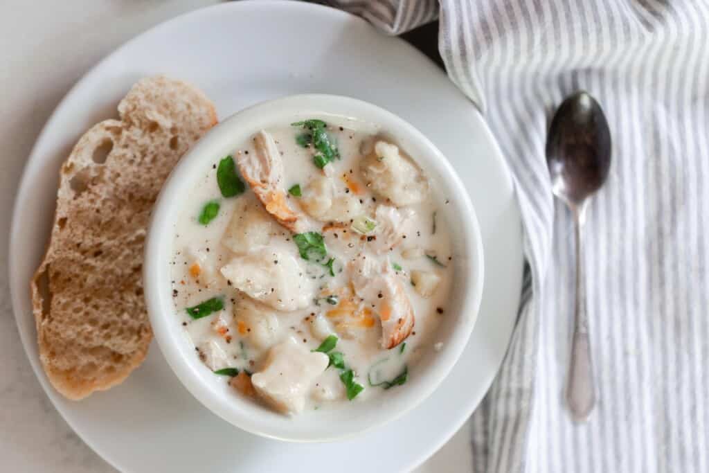 overhead photo of creamy homemade gnocchi soup in a white bowl on a white plate with a slice of sourdough bread to the left. A spoon sitting on a gray and white stripped napkin sits to the right