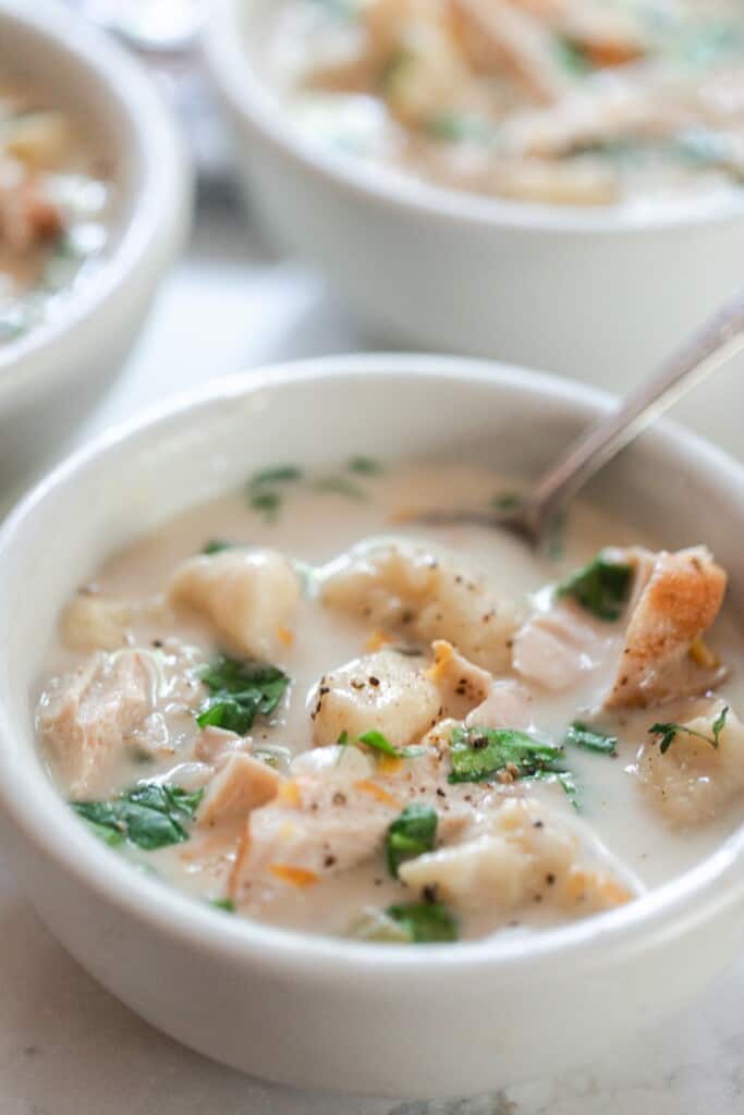 creamy gnocchi soup with chicken and herbs in a white bowl with a spoon in the soup. More bowls of soup in the background
