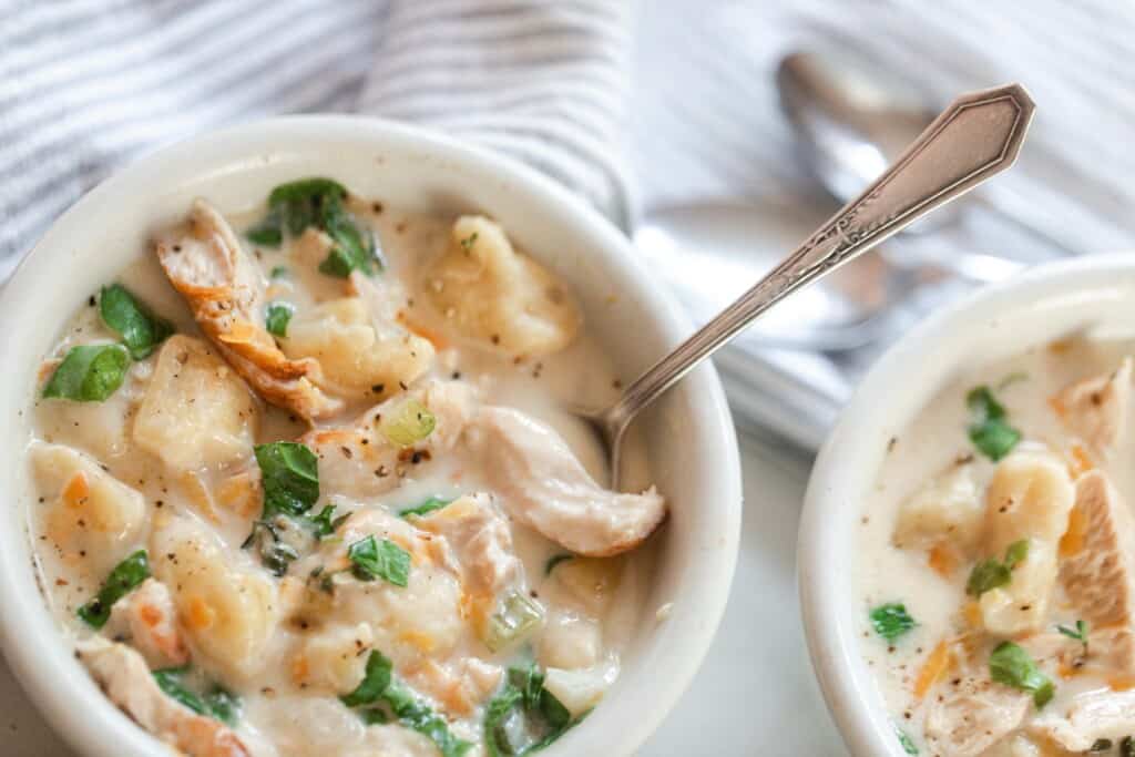 close up overhead photo of creamy gnocchi soup with chicken and herbs in a white bowl with a spoon. A gray and white towel with spoons sits directly behind the bowl.