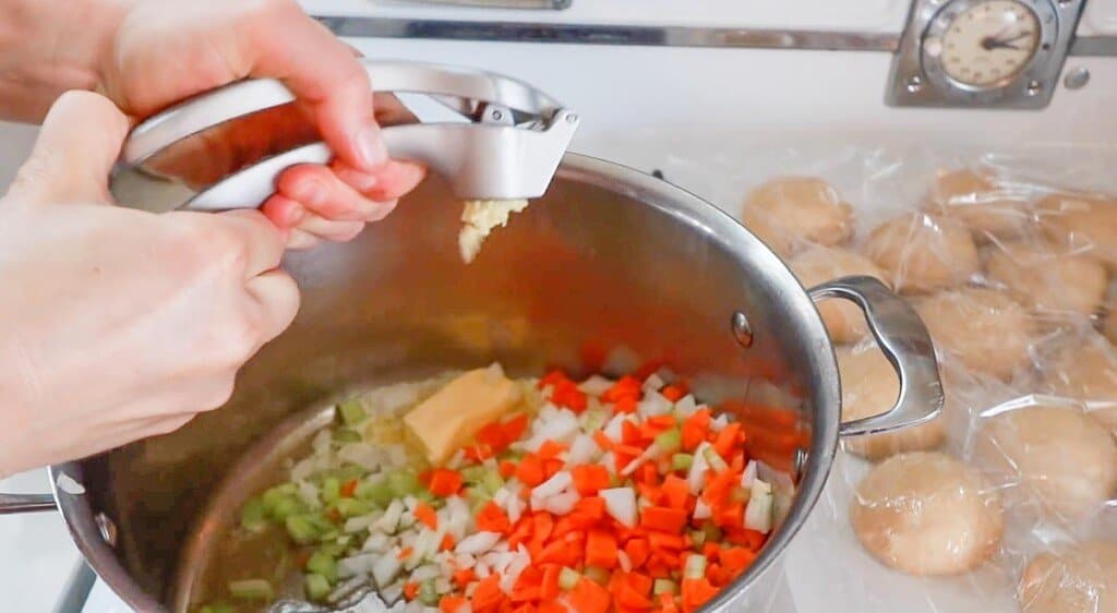 adding garlic with a garlic press to a pot of butter, diced carrots, celery and onions.