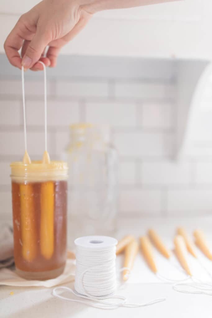 hand dipping cotton wick into a widemouth 1/2 pint jar with melted beeswax. A spool of wick is in front of the jar and more candles lay to the right