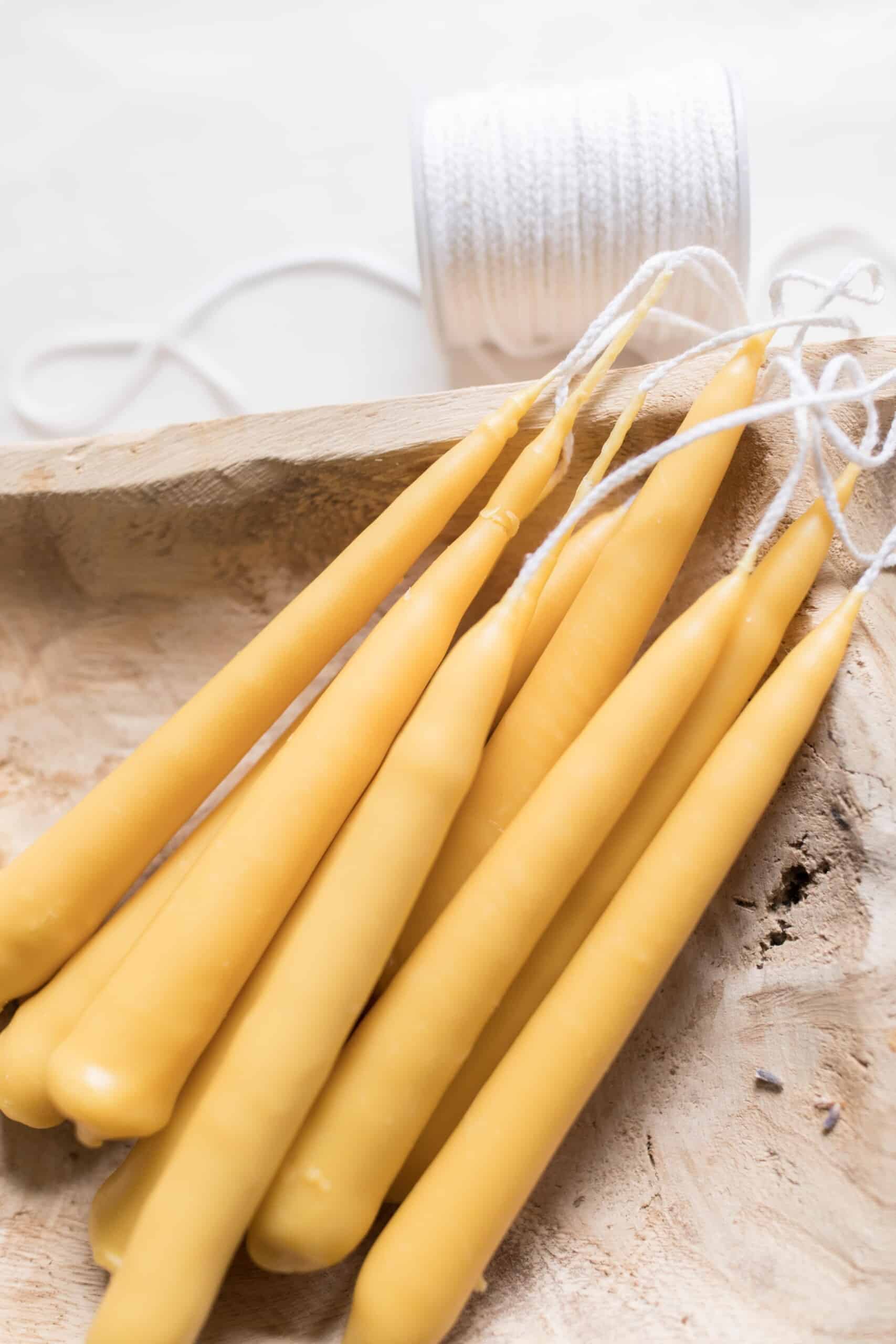 seven hand dipped tampered beeswax candles laying in a wooden bowl with a spool of wick behind the bowl