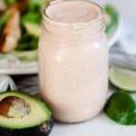 Mexican ranch dressing in a mason jar on a white counter with sliced avocados, limes around the jar and a salad in the background