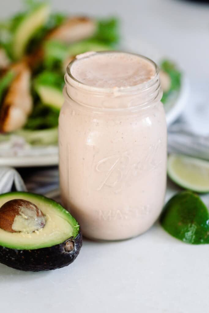 Mexican ranch dressing in a mason jar on a white counter with sliced avocados, limes around the jar and a salad in the background