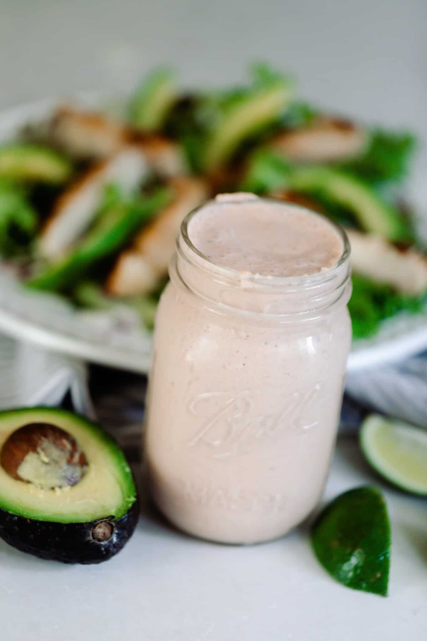 A mason jar of Mexican ranch dressing surrounding by avocados.