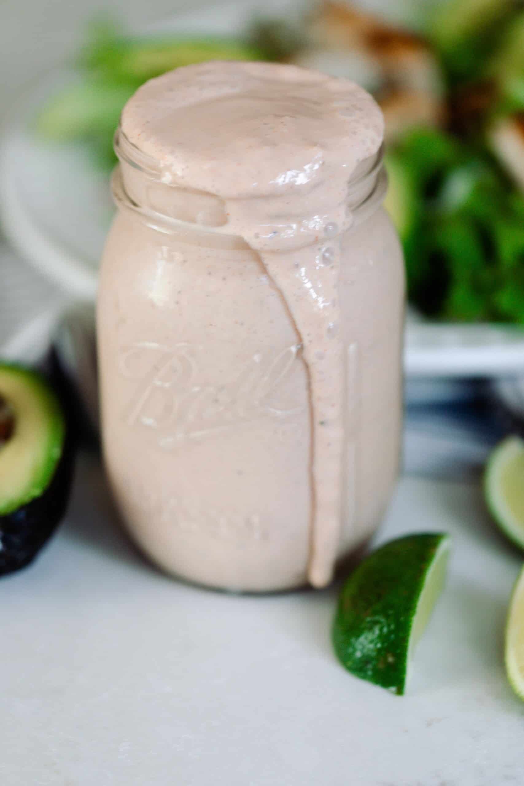 Mexican ranch dressing in a mason jar wit the dressing spilling over the top. Sliced limes and avocados lay on the counter around the jar of ranch