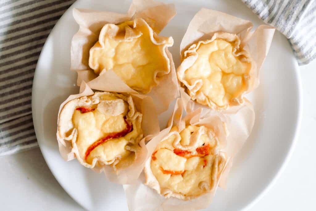 four mini quiches: two plain, two with roasted red pepper and potatoes, on a white plate on top a gray and white stripped towel