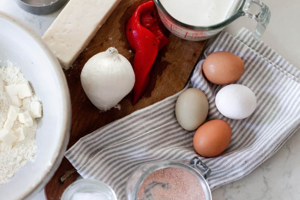 eggs on a gray and white stripped towel next to a cutting board with a roasted red pepper, whole onion, cheese, and milk on top of the cutting board. A large bowl of flour and cut but butter to the left