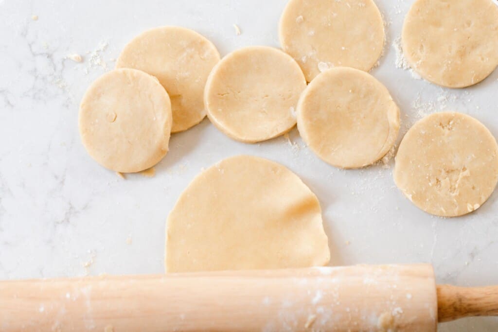 small biscuit sized pie crust on a white quarts countertop with a rolling pin rolling out the crust to make it thinner and slightly larger