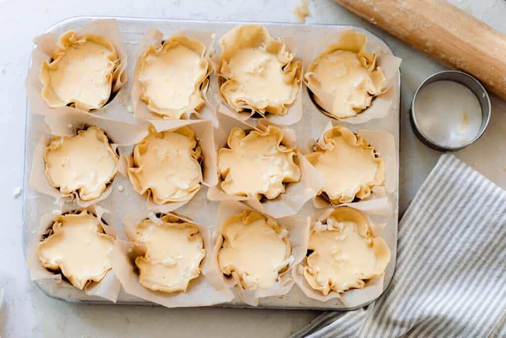 raw mini quiche in a parchment lined muffin tin with a rolling pin, biscuit cutter, and gray and white stripped towel to the right