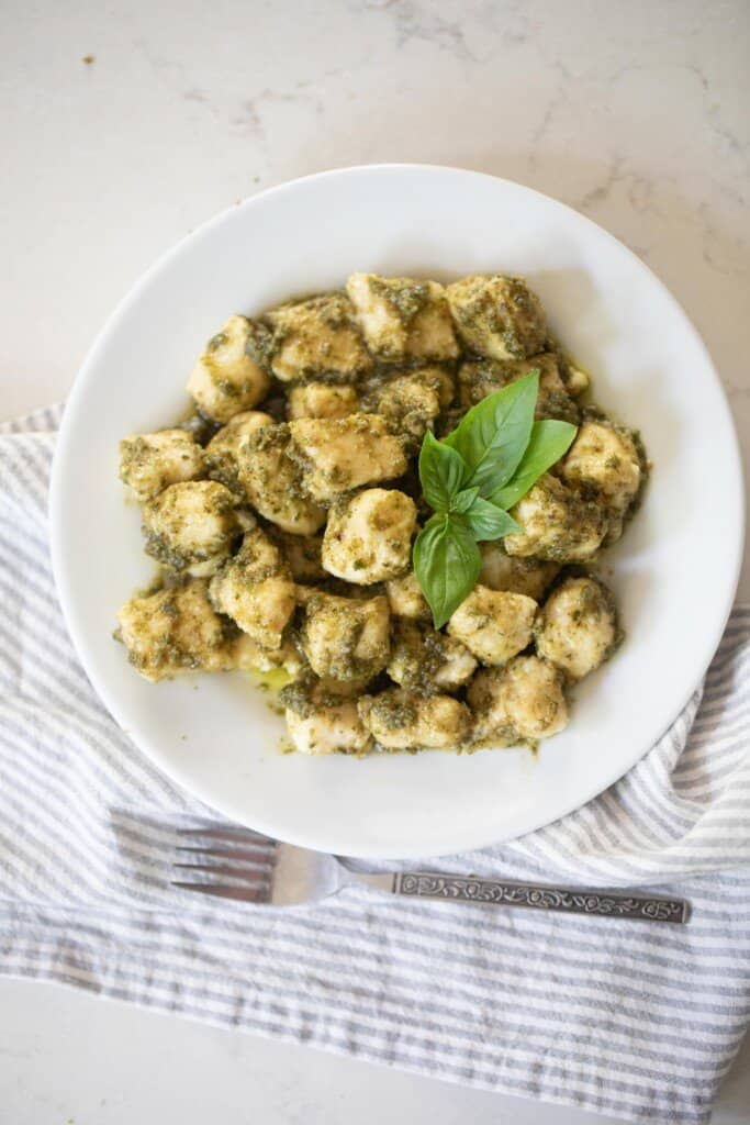 homemade pesto gnocchi garnished with five leaves of fresh basil on a white plate on top of a gray and white stripped napkin with a fork