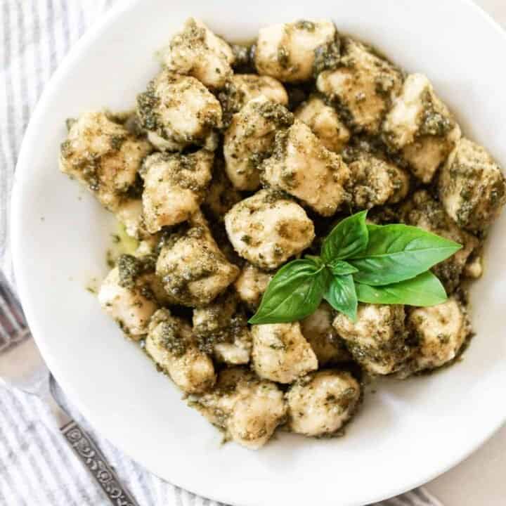 homemade gnocchi tossed in a fresh basil pesto and garnished with fresh basil leaves on a white plate with a blue ad white stripped napkin to the left