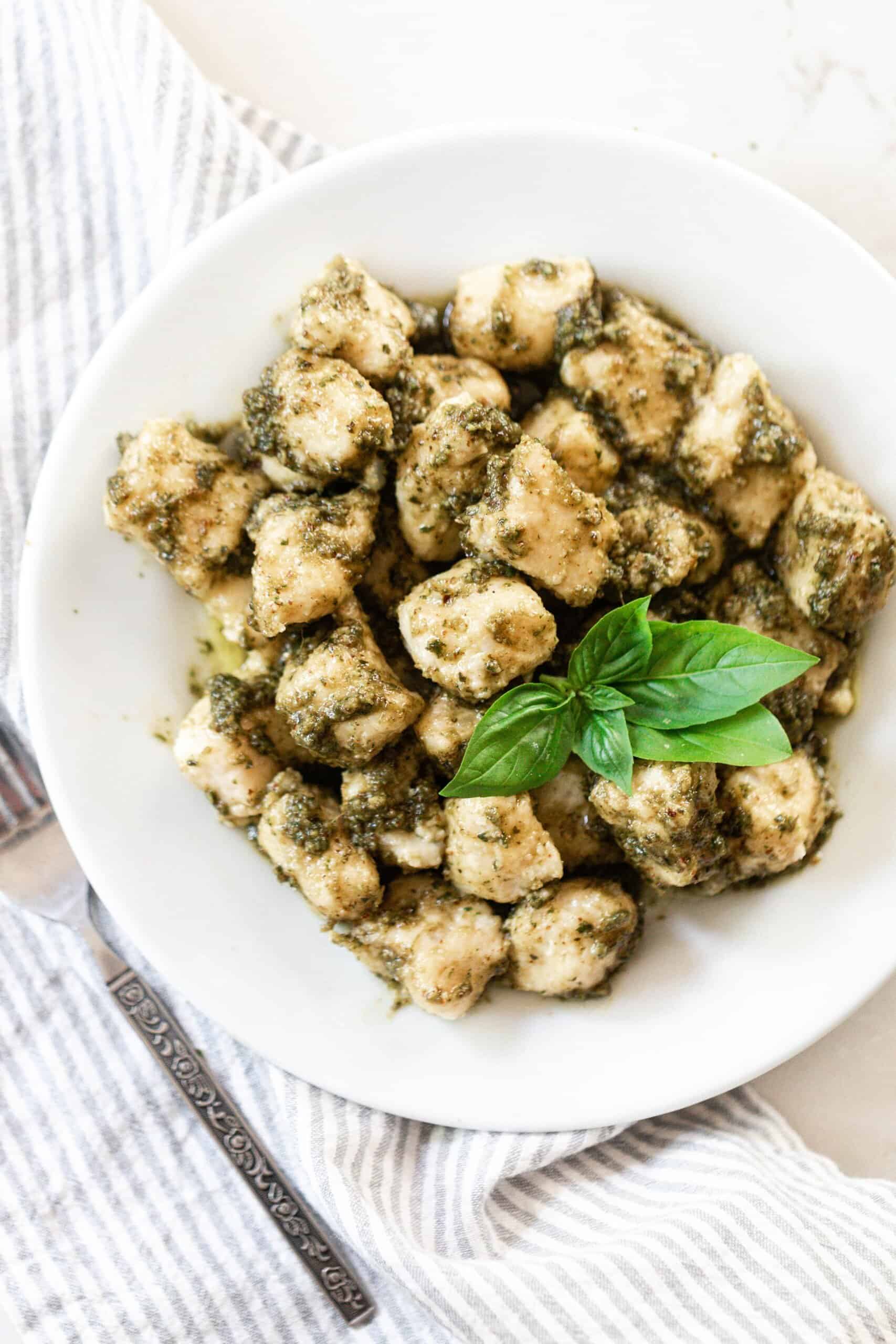 homemade gnocchi tossed in a fresh basil pesto and garnished with fresh basil leaves on a white plate with a blue ad white stripped napkin to the left