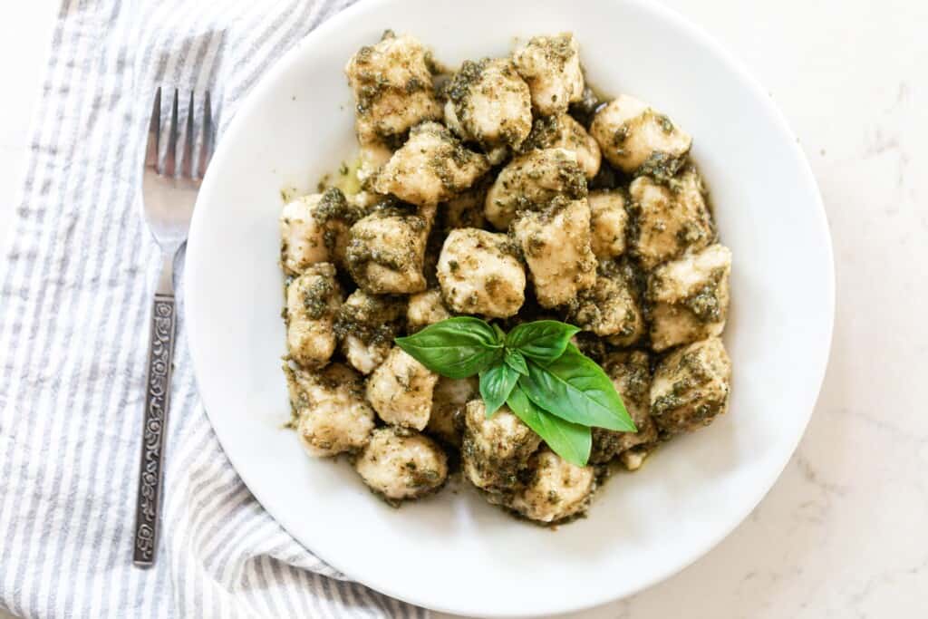 overhead photo of pesto gnocchi garnished with fresh basil leaves on a white plate with a white and blue stripped towel and a fork to the left