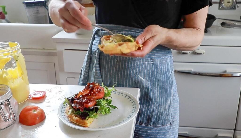 women wearing a blue stripped apron spreading mayo on a cheddar waffle half. The other waffle half is on a plate covered in tomatoes, lettuce, and bacon