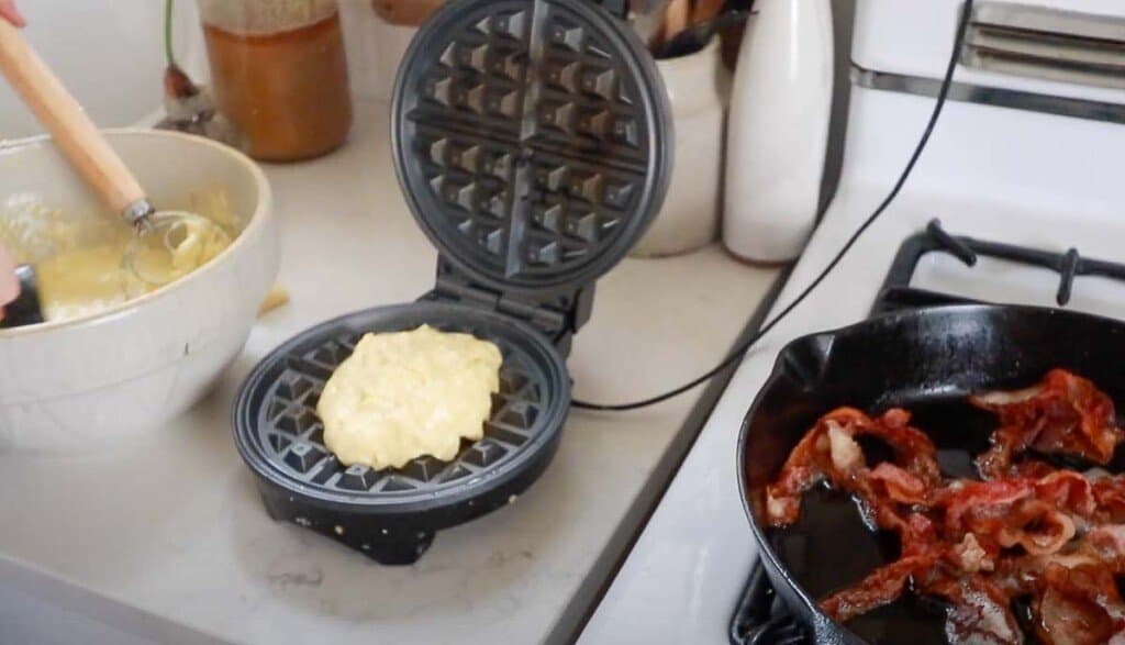 sourdough cheddar waffle batter in a waffle iron with a cast iron skillet full of bacon to the right.