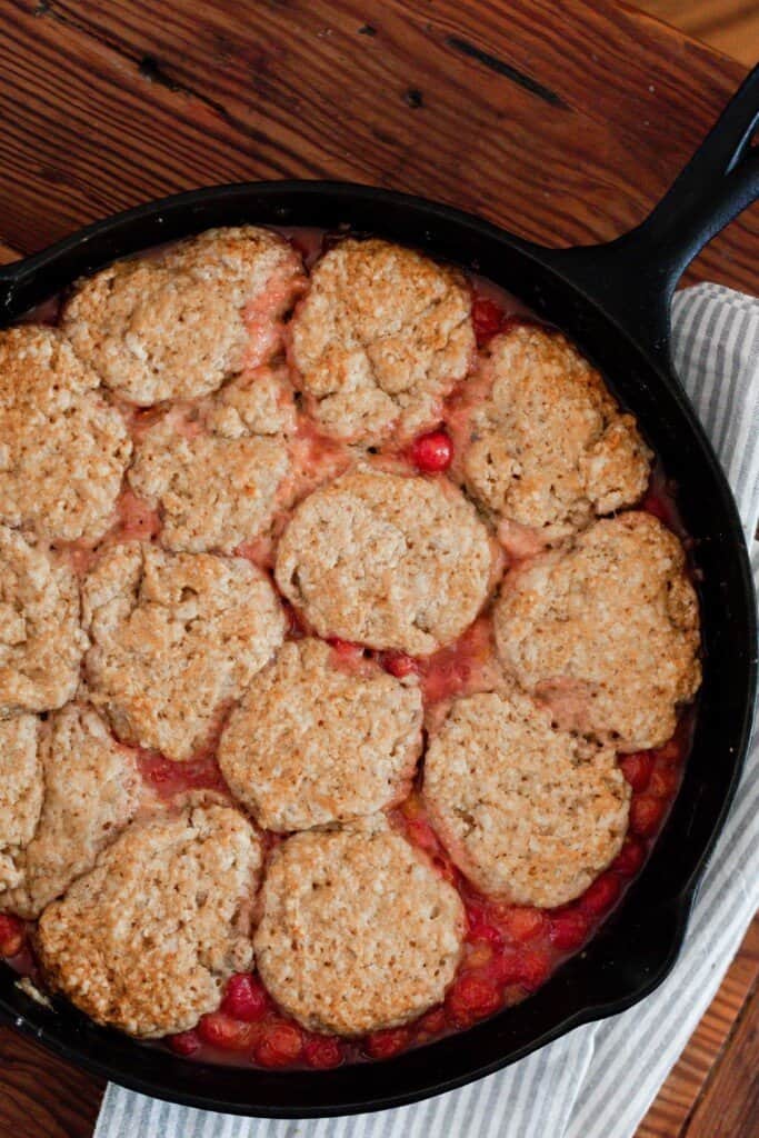 overhead photo of sourdough cherry cobbler in a cast iron skillet on top of a blue and white stripped towel laying on a wooden table