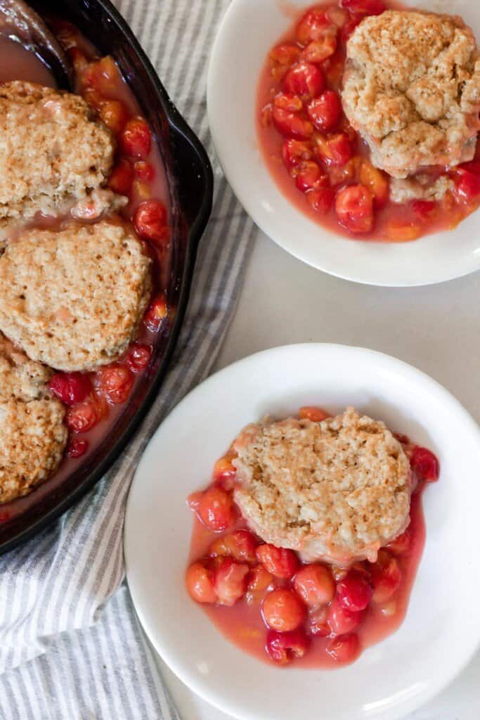 two white bowls of sourdough cherry cobbler on a white quarts countertop with a cast iron skillet with remaining cobbler to the left