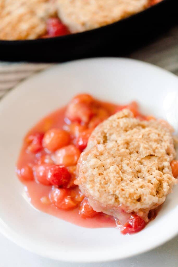 close up picture of a white bowl of sourdough cherry cobbler with a cast iron skillet of cobbler in the background