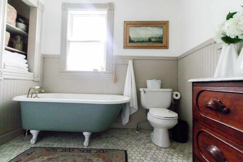 blue green antique clawfoot tub in a 1800s farmhouse bathroom. A toilet with art hanging above it sits to the right of the tub