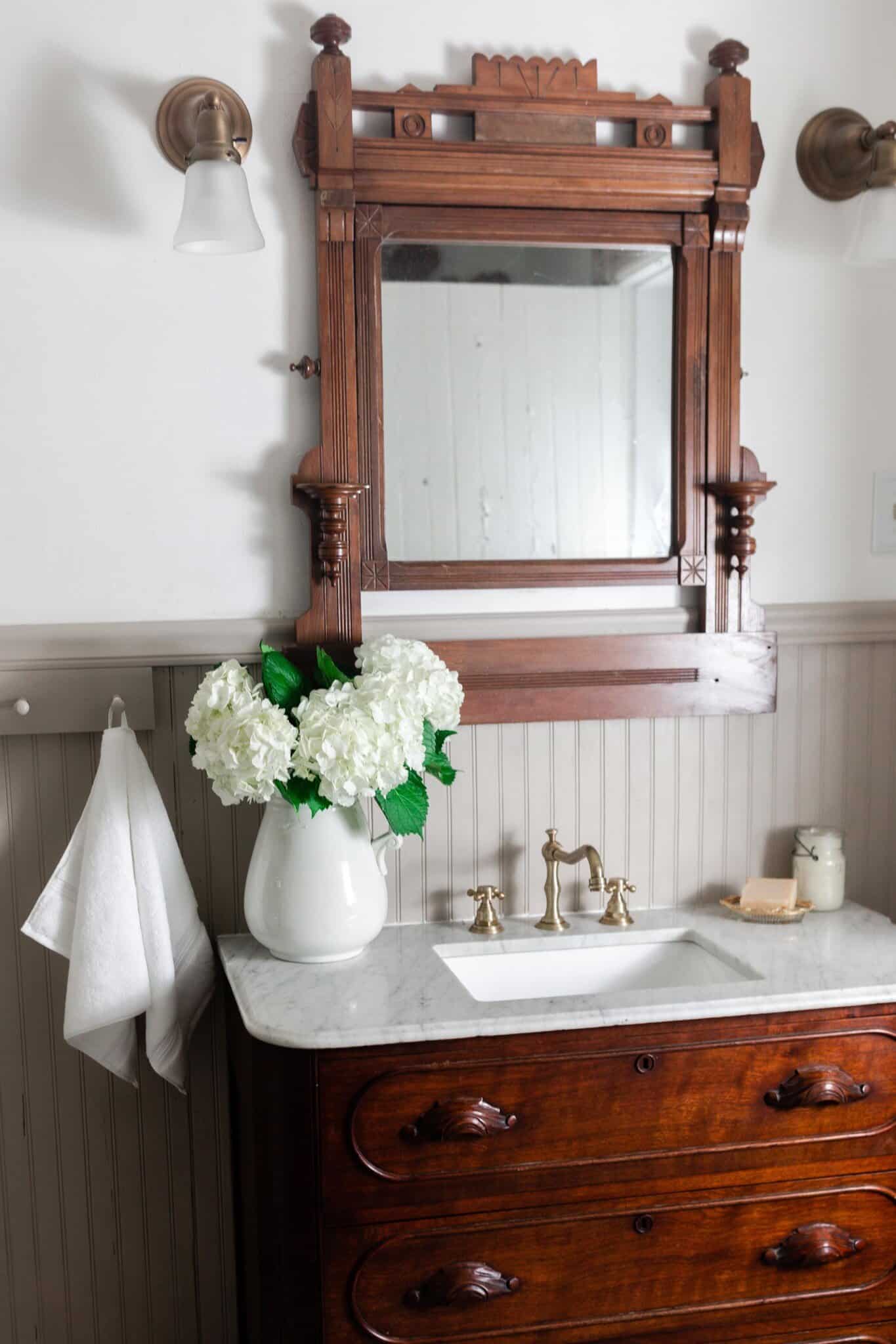 wood antique dresser with a marble top and sink with a vase of hydrangeas.A wooden antique mirror hands above the vanity with lights on each side of the mirror