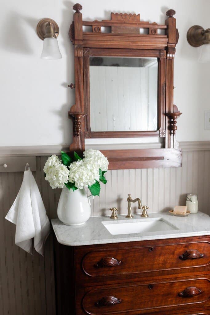 wood antique dresser with a marble top and sink  with a vase of hydrangeas.A wooden antique mirror hands above the vanity with lights on each side of the mirror 