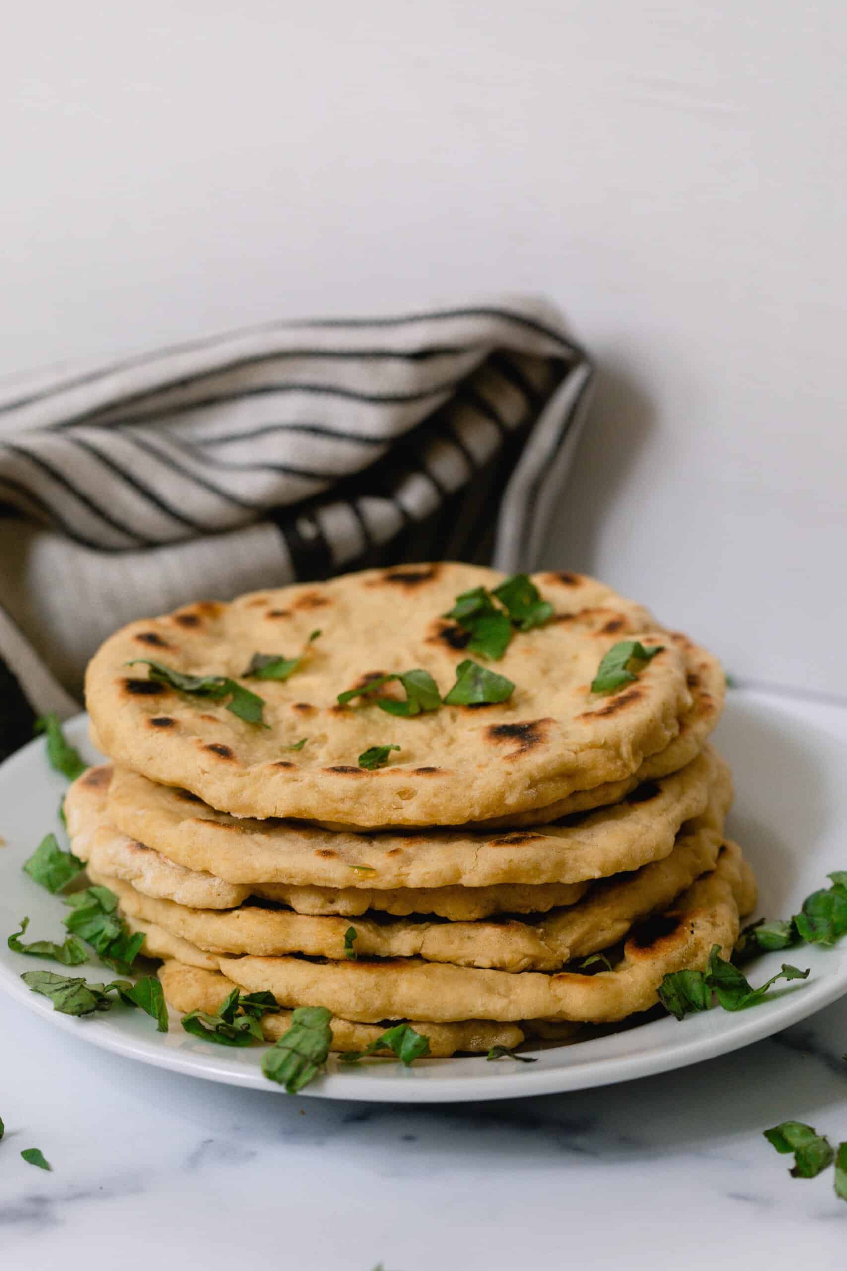 einkorn flat bread stacked with herbs sprinkled over on a white plate with a gray and black stripped tea towel in the background