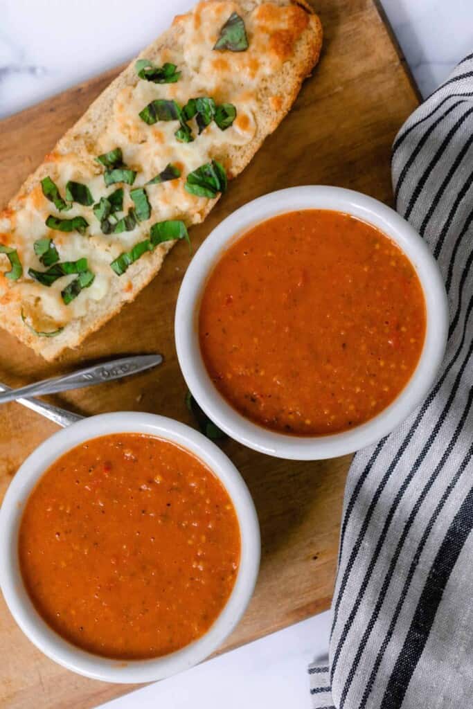 two bowls of homemade tomato soup is white bowls on a wood cutting board with a white and black towel to the right and a cheesy herb bread behind the bowls