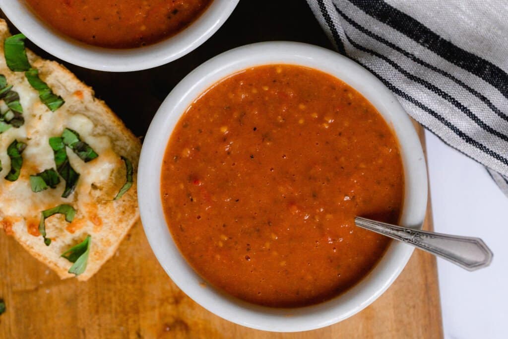 overhead photo of fresh tomato soup in a white bowl on a cutting board with herb and cheese bread to the left