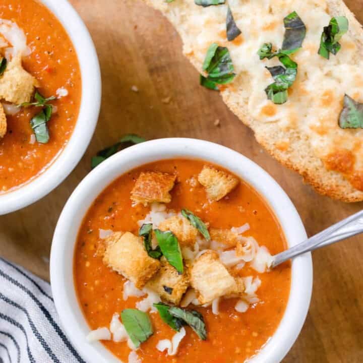 two bowls of tomato soup from scratch with fresh tomatoes topped with fresh basil, croutons, and cheese on a wood cutting board with a loaf of cheesy bread and a white and black stripped towel to the left