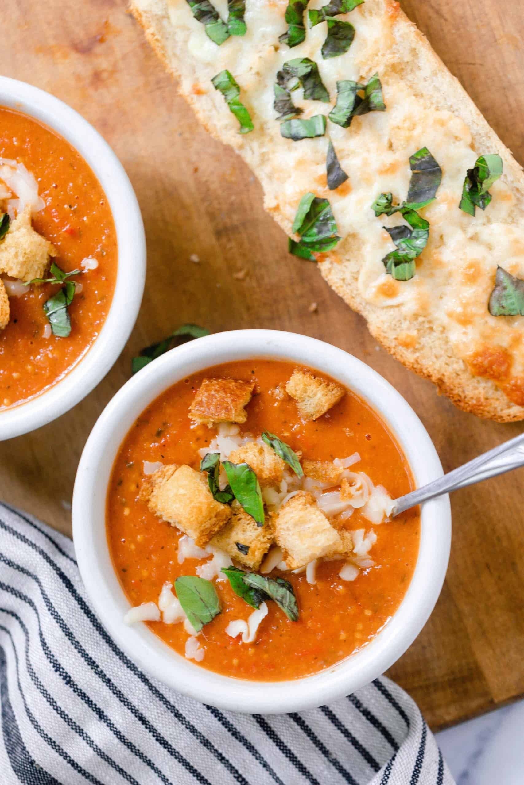 two bowls of tomato soup from scratch with fresh tomatoes topped with fresh basil, croutons, and cheese on a wood cutting board with a loaf of cheesy bread and a white and black stripped towel to the left