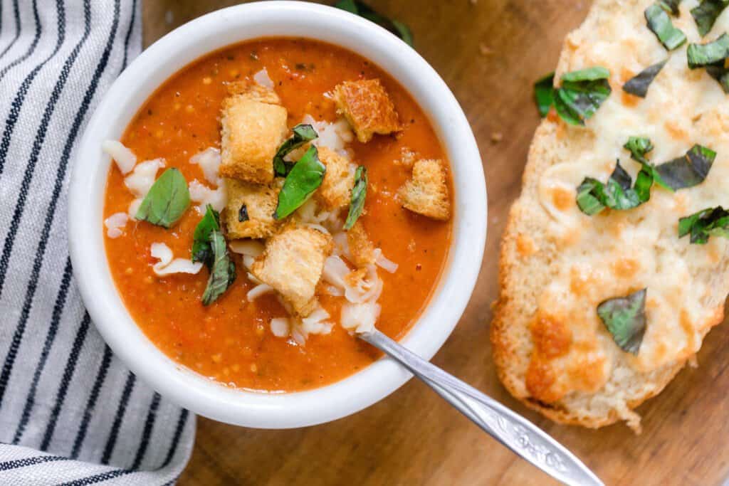 overhead photo of tomato soup made from fresh tomatoes topped with bacon, cheese, fresh basil and croutons on a wood cutting board with cheesy bread to the right