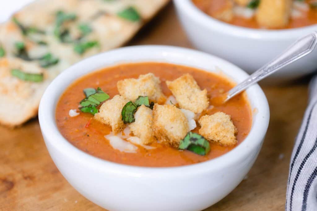 close up picture of a white bowl full of tomato soup with homemade croutons, fresh basil, and cheese in a bowl on a wood cutting board