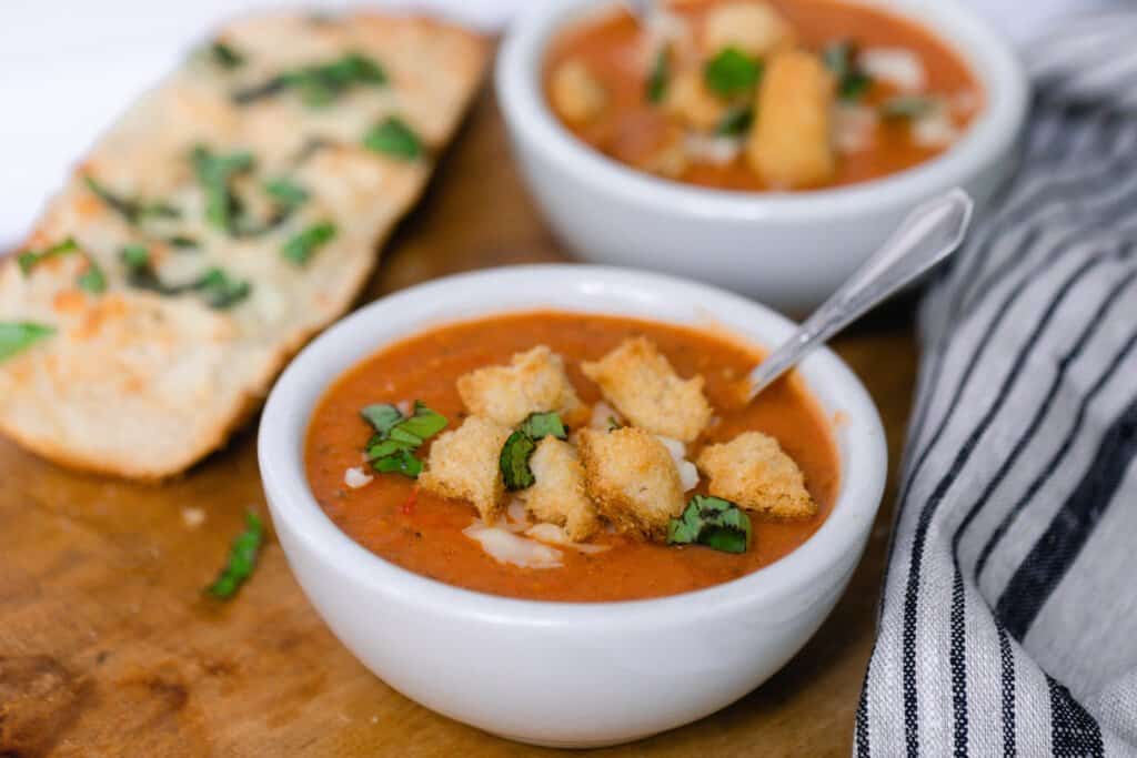 two bowls of tomato soup topped with herbs, cheese, and croutons on a wood cutting board with a black and gray stripped towel to the right