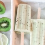 two kiwi lime popsicles in a white dish with ice. Halved kiwis and a lime sit to the left of the dish