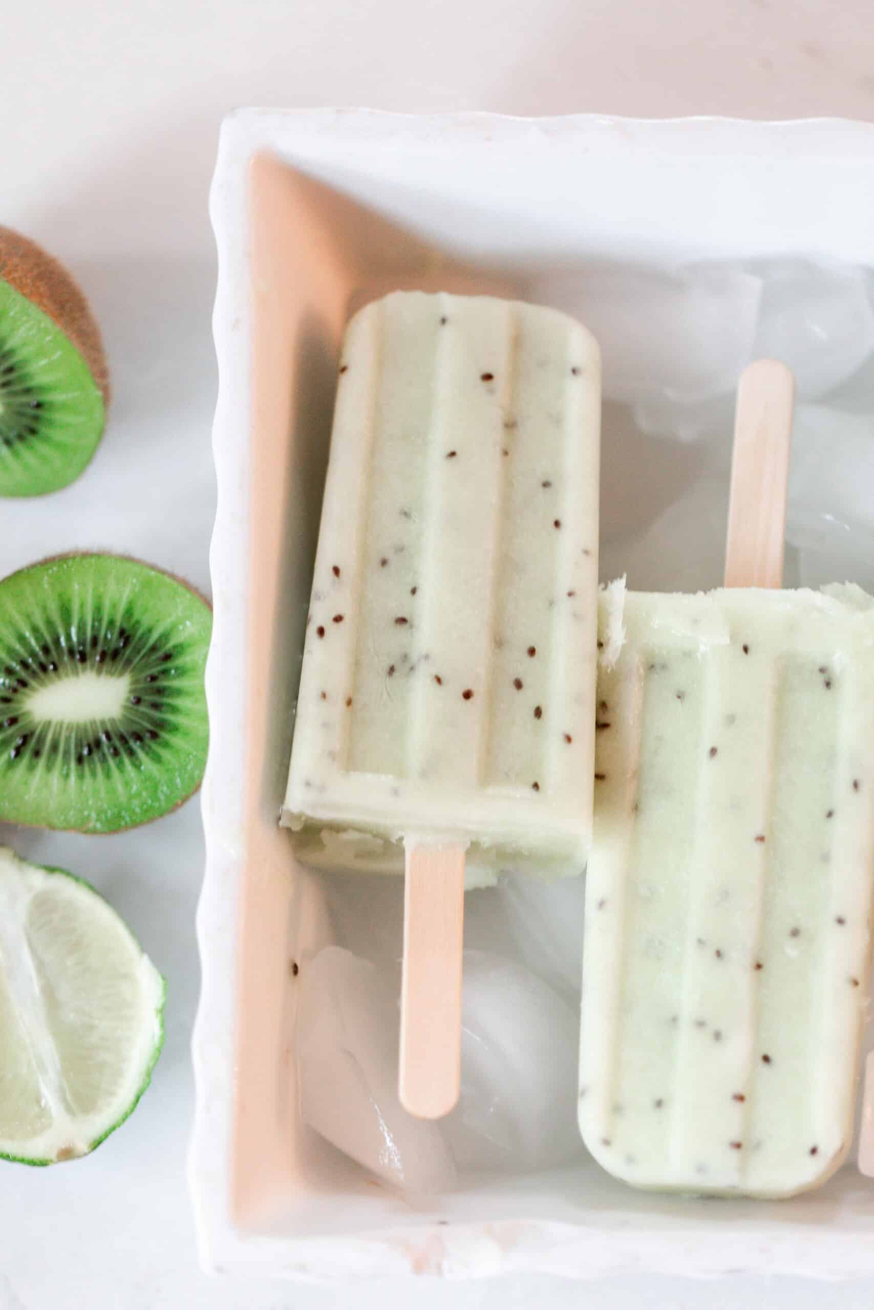 two kiwi lime popsicles in a white dish with ice. Halved kiwis and a lime sit to the left of the dish