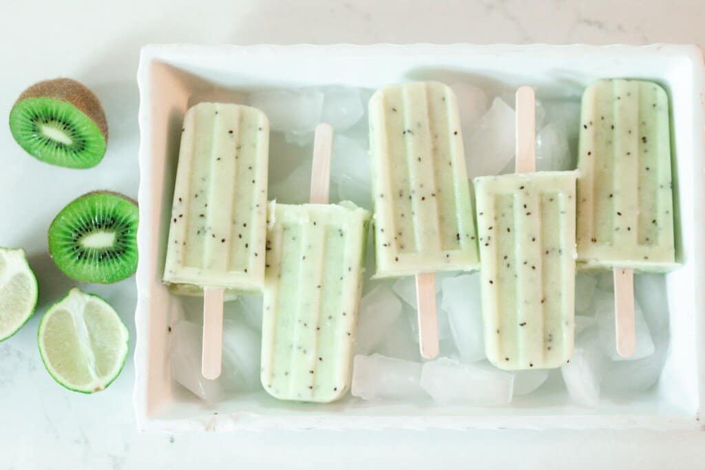 five kiwi lime popsicles laying on a bed of ice in a white dish with sliced kiwi and lime to the left