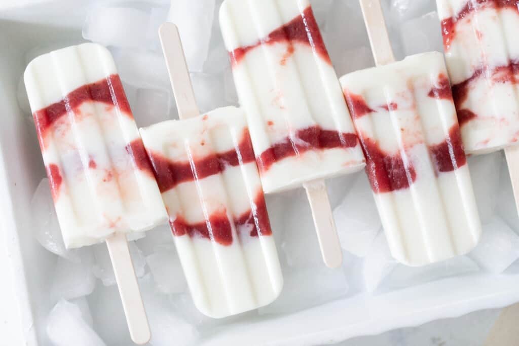 strawberry yogurt popsicles laying on a dish of ice. The popsicles are made with layers of yogurt with layers of strawberry jam