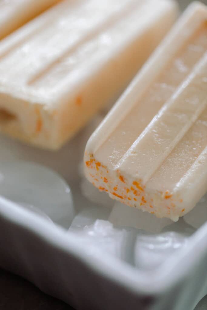 close up picture of light orange creamy popsicles with orange zest at the top of the popsicles that are laying on ice