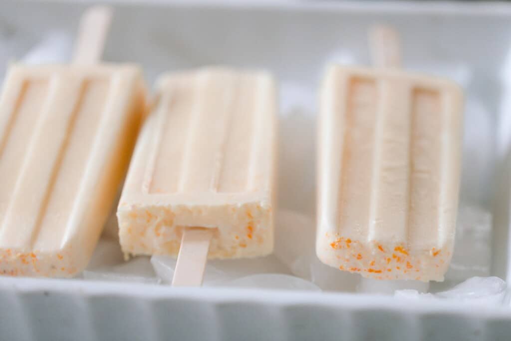 three orange creamsicle popsicles laying on ice in a white baking dish