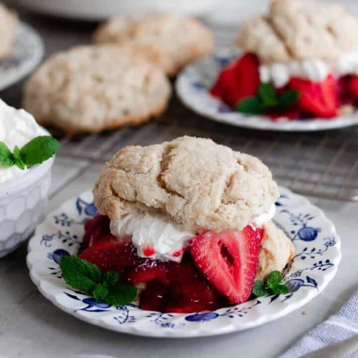 two strawberry shortcakes with whipped cream and strawberries layered between the biscuits on a white and blue plates with extra biscuits and whipped cream behind the plates