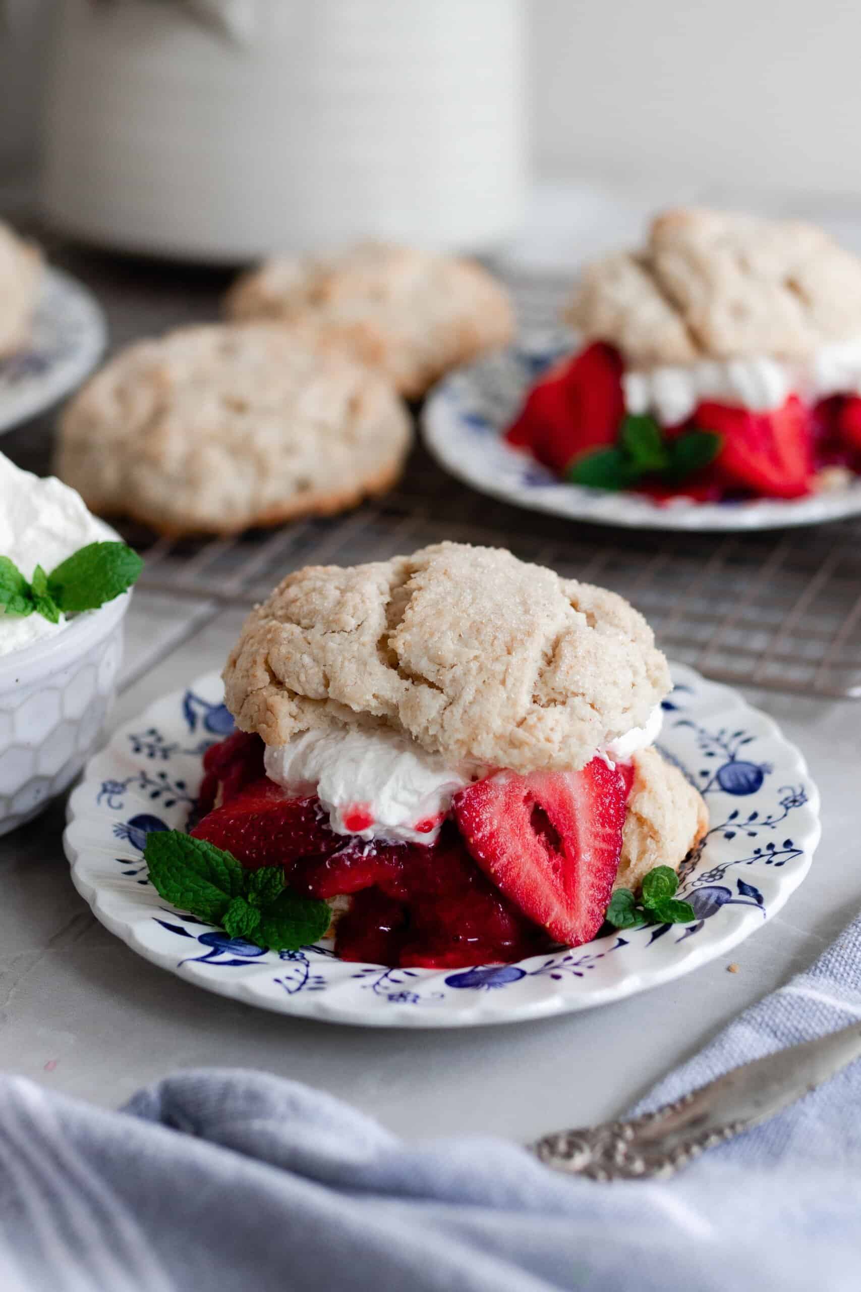 two strawberry shortcakes with whipped cream and strawberries layered between the biscuits on a white and blue plates with extra biscuits and whipped cream behind the plates